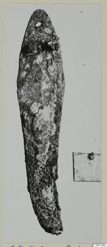 Corroded metal tool with a tapering blade and a small hole near the tip, photographed beside a small label card