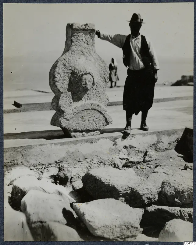 Man in a hat resting his hand on a weathered carved stone figure at the palace at Vouni.