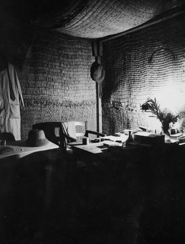Interior of a bast house at Mersinaki with a reed-woven wall, a table with a hat, a chair and a vase of leaves.