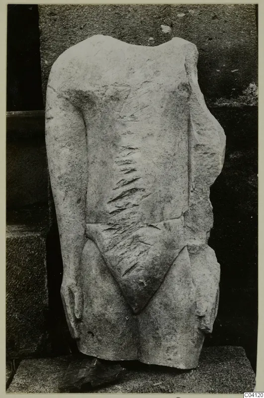 Limestone sculpture of a headless, armless standing figure torso, shown against a dark stone wall.