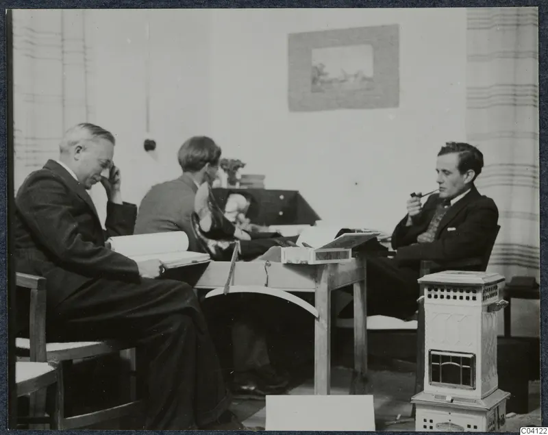 Three men seated in a room in Nicosia, with a table and books; one reads and one smokes a pipe.
