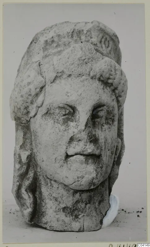 Limestone female head from Soli, shown frontally on a plain background, nose and parts of the headdress broken.