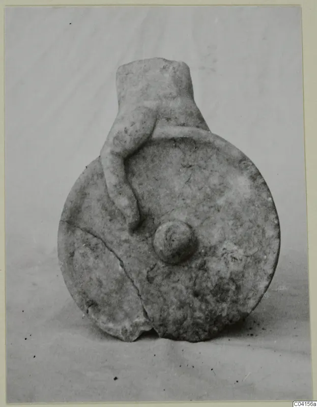 Carved stone object: a round flat disc with a cylindrical neck, with a small figure clinging to its side.