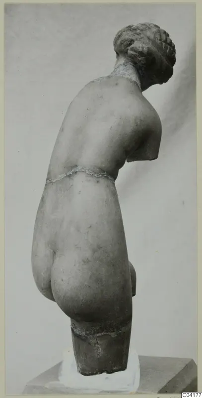 Back view of a marble statue of a nude figure with the head bowed and hair gathered in a coiled bun, the arms broken off.