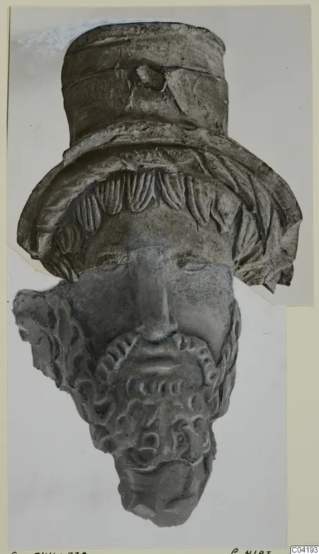 Terracotta head fragment with a tall cylindrical headdress and a curled beard, photographed on a pale card mount