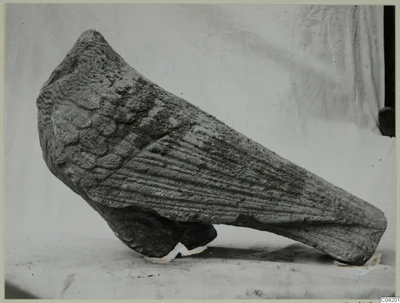 Stone fragment from Soli, tapering, with overlapping scale-like carving on one side and horizontal ridges on the other.