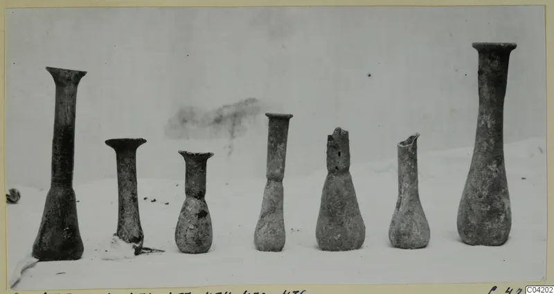 Seven blown glass vessels with narrow necks standing in a row on a pale surface.