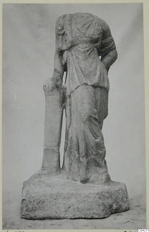 Headless marble statue of a standing draped female figure beside a tall narrow support, on a rough stone base