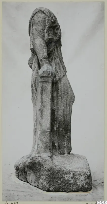 Stone statue of a draped standing figure on a rough block base, photographed against a plain grey backdrop.