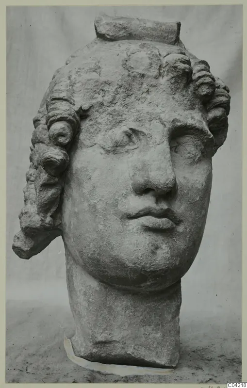 Carved stone head of a figure with curled locks and a raised band across the top, photographed against a grey backdrop. Soli.
