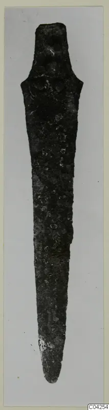 Corroded bronze blade with a short tang, from Milia, photographed upright against a grey background