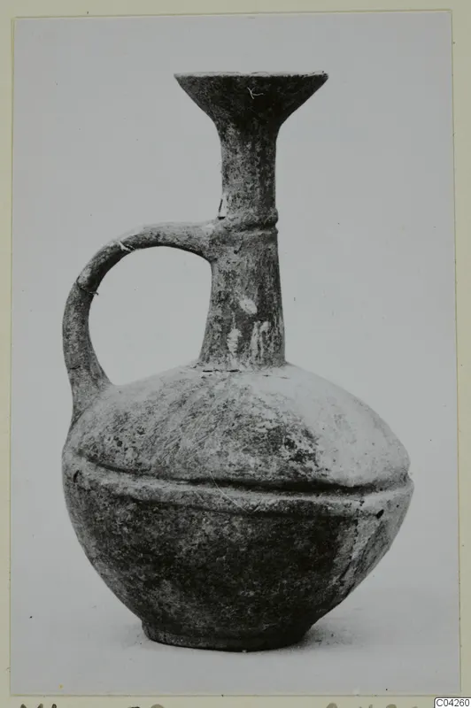 Base-ring style jug with a single loop handle and a flaring trefoil-like rim on a tall neck, photographed against a plain background.