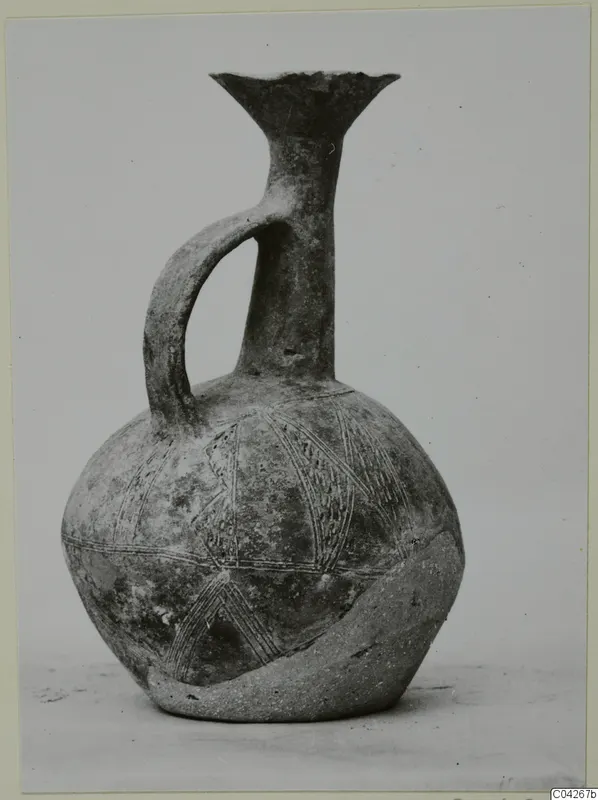 Black-slipped jug with a trefoil-like flared mouth, single strap handle and incised linear decoration, photographed against a grey backdrop.