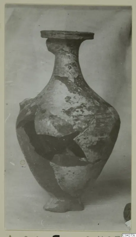 Pale jar with a flat rim, narrow neck and broken body, photographed alone against a plain backdrop.