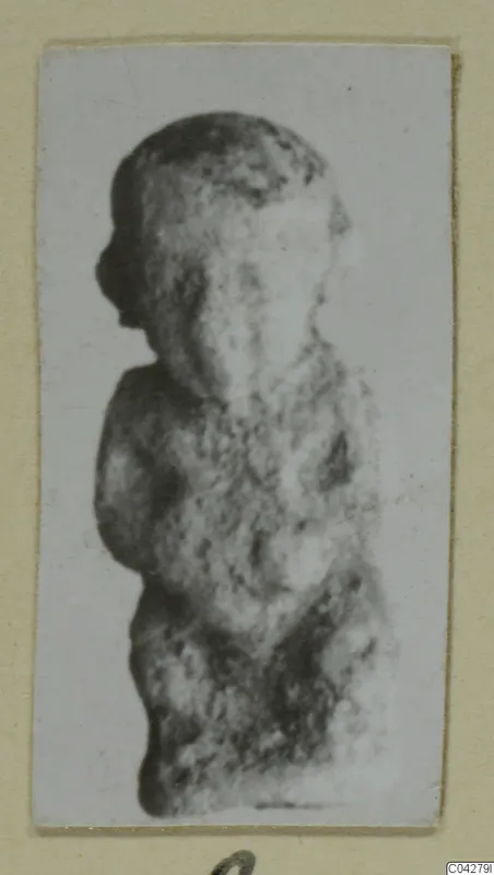 Small worn figure of a standing human figure, seen in side view, in a photograph mounted on a cream card