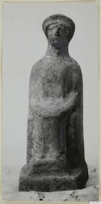 Terracotta figurine of a standing figure with a cap-like headdress and folded arms, on a square base.