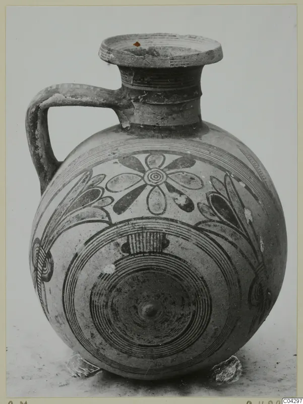 Round-bodied jug with a single handle and painted concentric circles and flower motifs in dark paint on a pale surface.