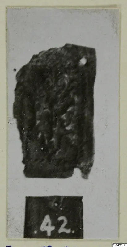 Fragment of tarnished silver sheet with raised scroll-like pattern, shown with a number label below