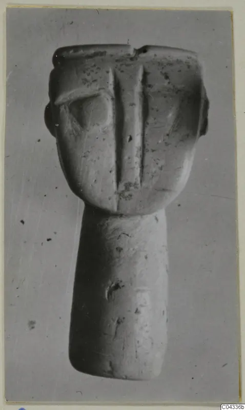 Stone figurine with a flat-topped head, a central ridge down the face and a long cylindrical body, photographed against a grey background.