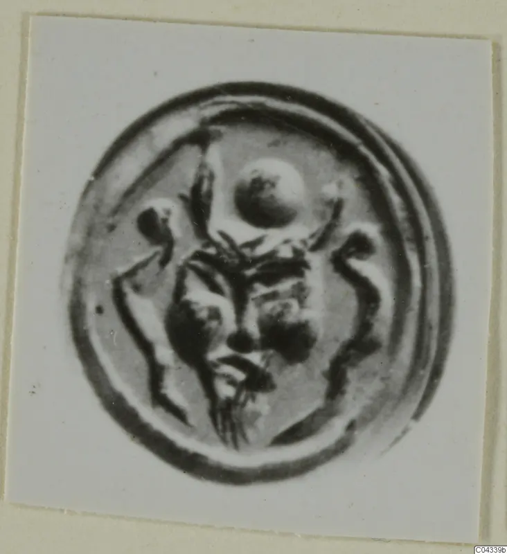 Round seal or gem in a circular frame, with a raised central motif of a face-like shape and a round element above it