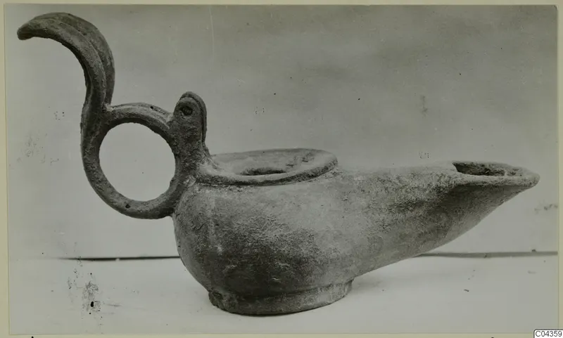 Metal oil lamp with a ring handle topped by a curled projection, spout to the right, shown in side view.