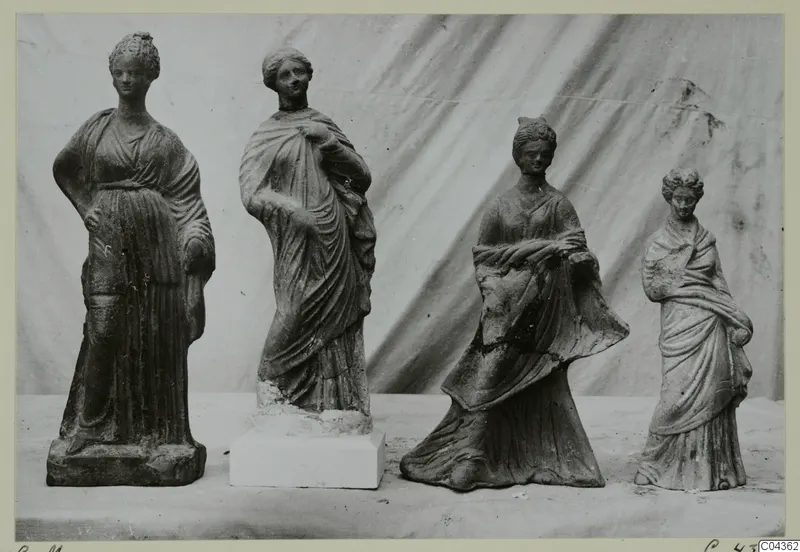 Four standing female terracotta figurines in draped robes, set in a row on a table against a draped cloth.
