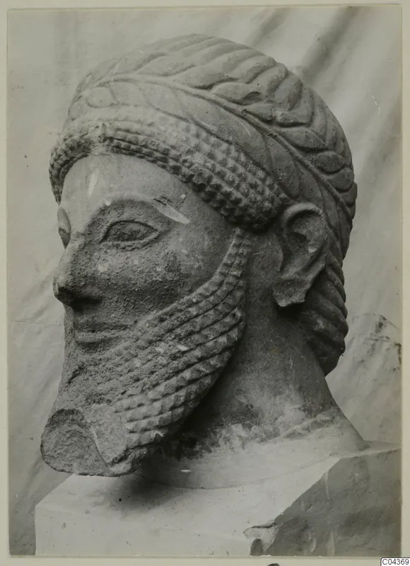 Limestone head of a bearded male figure with a leafy band around the crown, shown on a plinth against a draped backdrop.