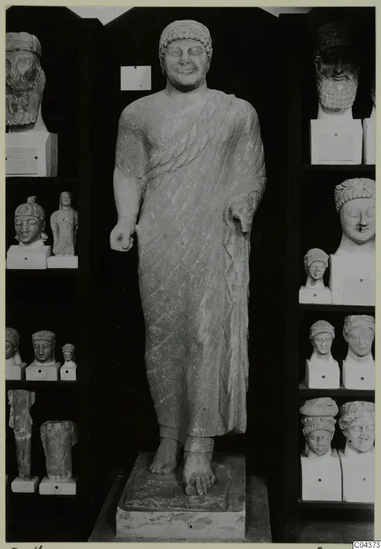 Standing limestone statue of a draped male figure, displayed on a base between shelves of heads and busts