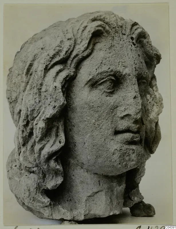 Carved stone male head with long wavy hair, shown in three-quarter view against a plain background.