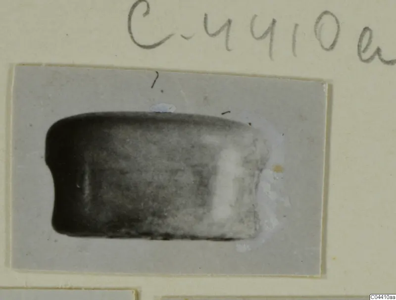 Small photograph of a dark stone seal or gem-like object, roughly rectangular with rounded ends, mounted on a card