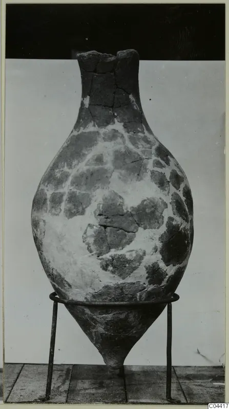 Large bulbous vessel with a narrow neck and broken rim, standing on a metal ring stand against a plain backdrop