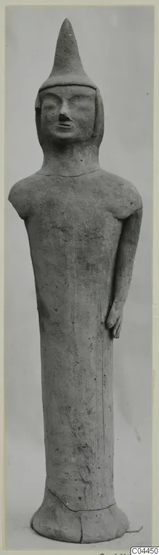 Standing terracotta figure with a tall pointed headdress and elongated body, photographed against a grey backdrop.