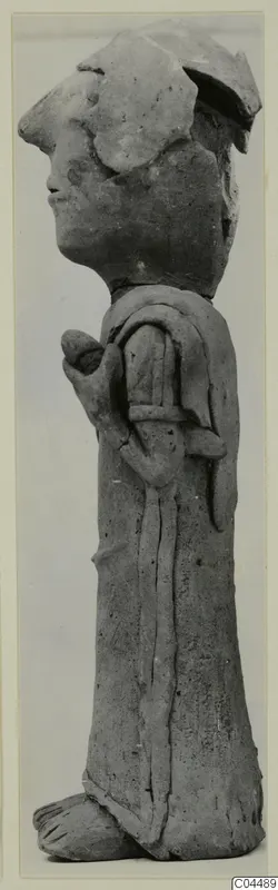 Side view of a terracotta standing figurine with a flat headdress and one arm bent across its body.