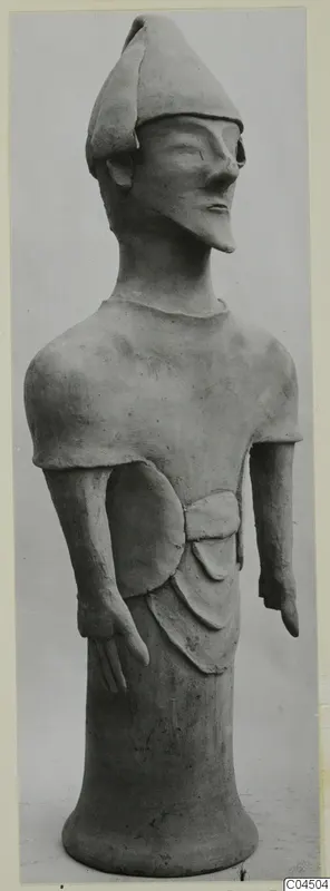 Standing terracotta figure wearing a pointed cap, arms at its sides, shown against a plain grey background
