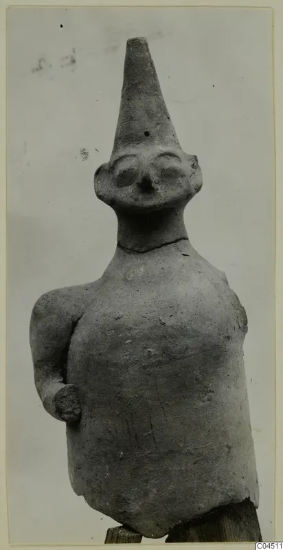 Terracotta figurine from Ayia Irini of a standing human figure with a tall conical head and one arm bent at the side.