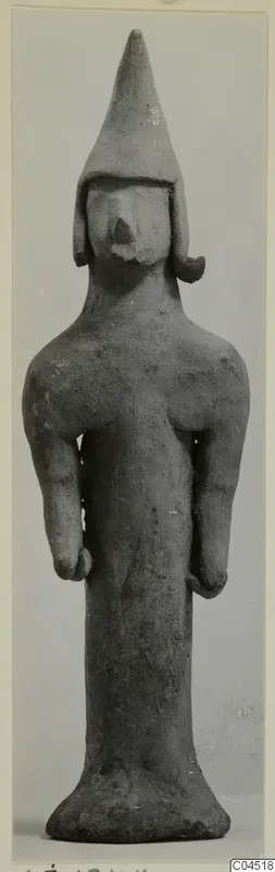 Terracotta figurine of a standing figure with a tall conical head covering and arms held at the sides.