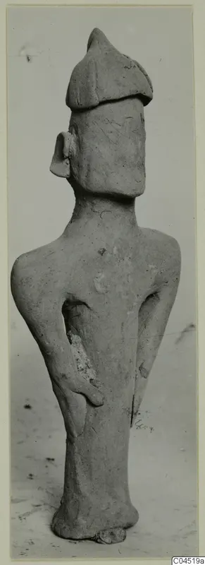 Terracotta figurine of a standing figure with a tall cap and bent arms, photographed against a plain grey background.