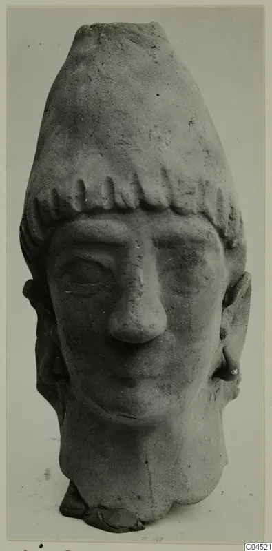 Terracotta head of a figure with a tall flat-topped headdress and fringe of curls, face with broad nose and downcast eyes.