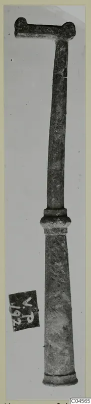 Long bronze tool with a T-shaped top and tapering shaft, photographed upright against a plain background.