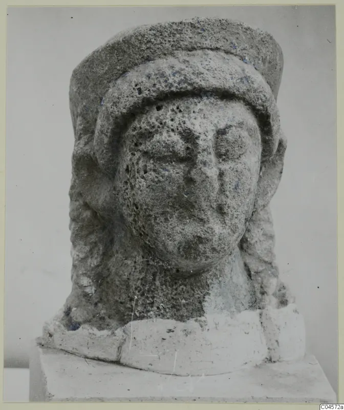 Limestone head sculpture from Arsos, with a tall cylindrical headdress and long locks of hair.
