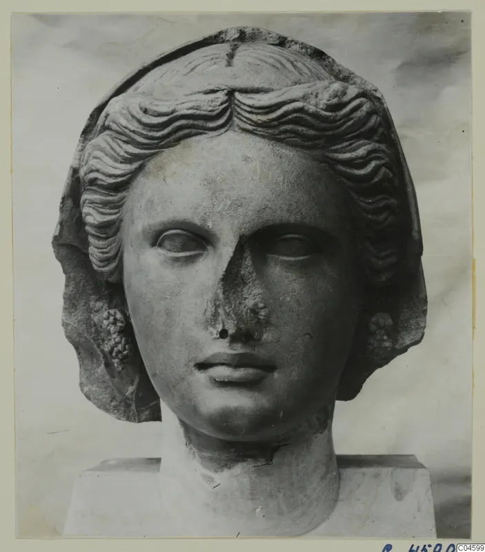 Stone head of a woman with wavy hair and a veil, nose damaged, set on a plinth