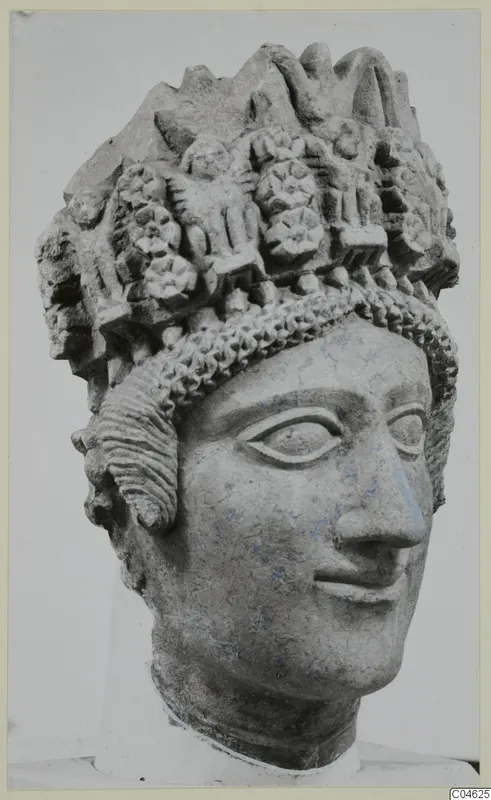 Carved limestone head of a figure wearing a tall headdress of rosettes and rounded motifs, photographed against a grey backdrop.
