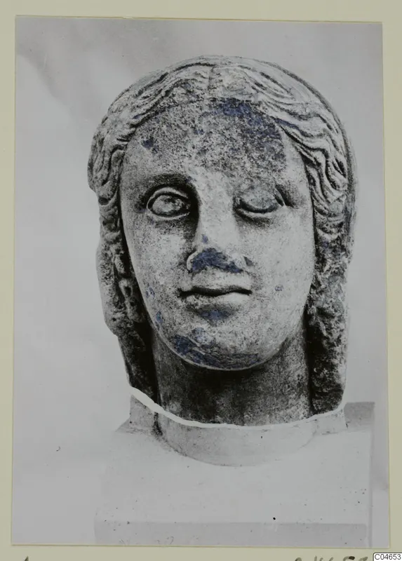 Stone statue head from Arsos, shown frontally on a block, with one eye partly closed and blue-grey staining on the face.