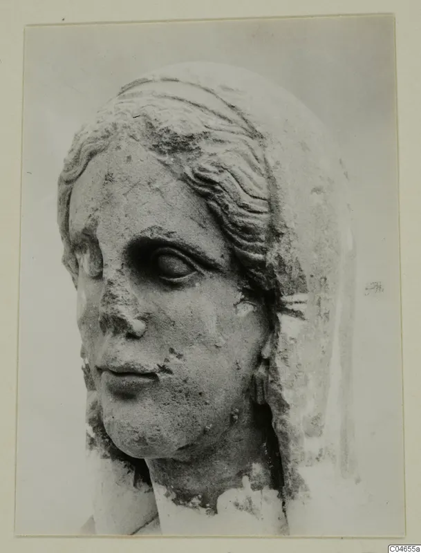 Stone statue head from Arsos, a female face with wavy hair over the brow, with the nose and surface worn away.