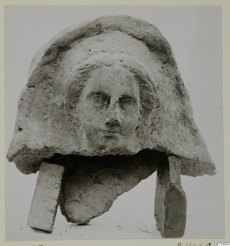 Carved stone head fragment with a hood-like headdress and worn face, propped on two rectangular blocks