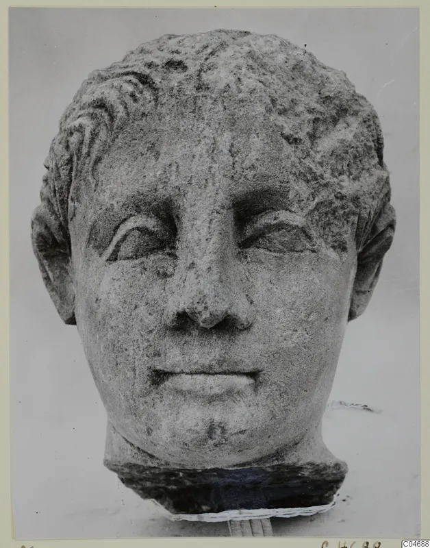 Carved stone head with short wavy hair and a worn face, set on a dark block against a pale background