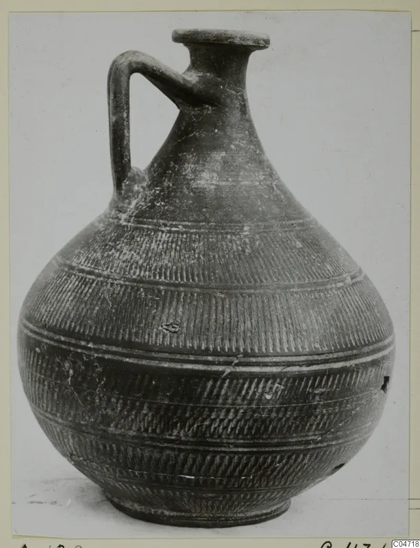 Dark pottery jug with a single loop handle and incised bands, photographed against a pale background, from Amathus