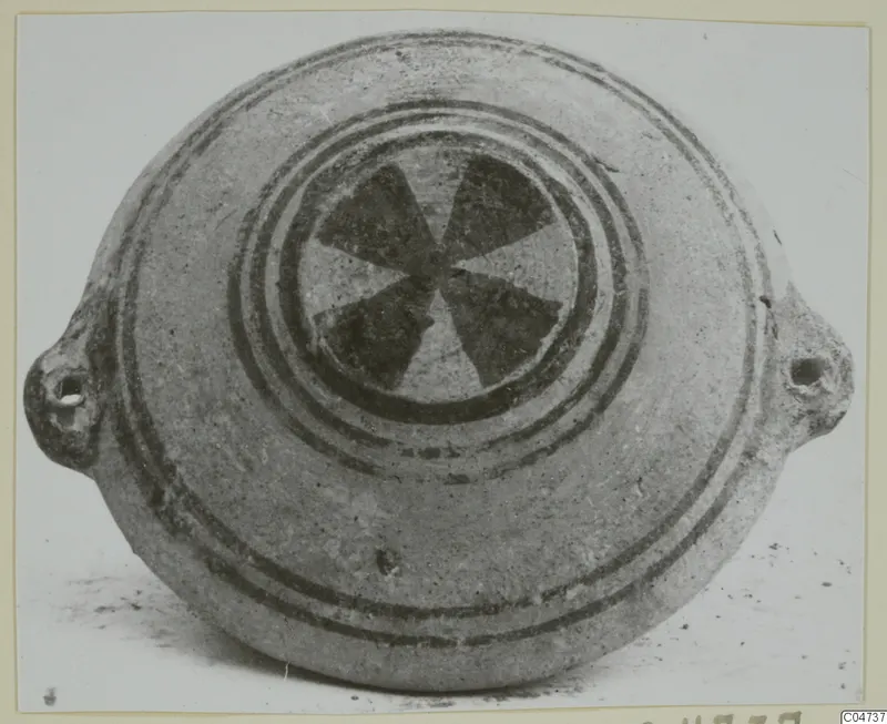 Pottery White Painted II: a flat, disc-shaped vessel with two pierced lugs and a dark six-spoked motif at the centre of its top