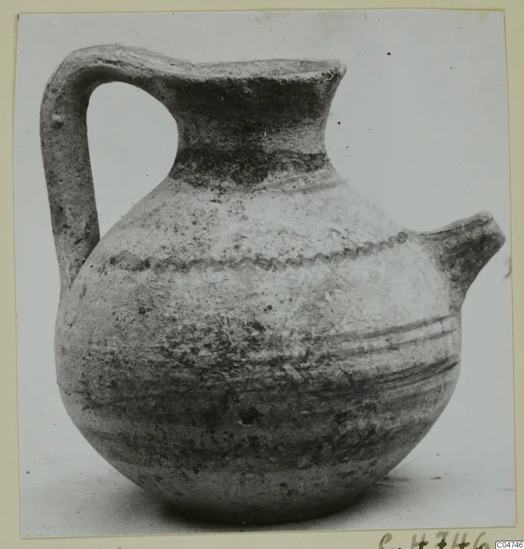 Jug with a strap handle, a trefoil-like spout and a wavy incised band around the shoulder, photographed on a plain surface.