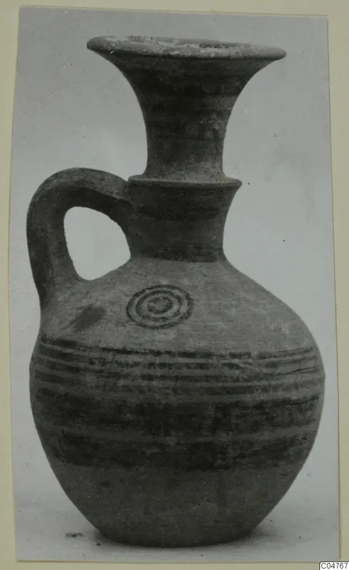 Pottery juglet with a trefoil-like flared neck, single loop handle, and dark painted bands and concentric circle on a buff body.